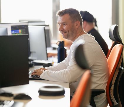 Mann im weißen Hemd lächelt, während er in einem Büro mit orangefarbenen Stühlen an einem Computer arbeitet. Eine weitere Person sitzt in der Nähe.