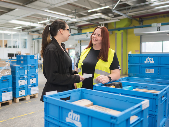 Две служителки в разпределителния център стоят между сини складови кутии и обсъждат нещо помежду си. На заден план се виждат още кутии на палети. Служителките изглеждат мотивирани и весели.