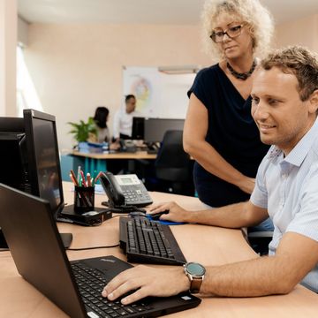 Мъж работи на лаптоп на бюро, докато жена стои до него и наблюдава. Двама души се намират на заден план в офисна обстановка.