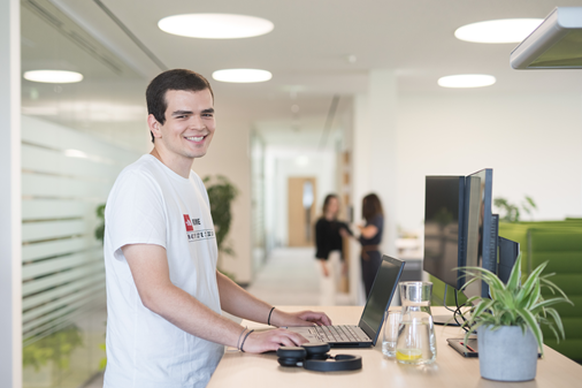 Ein Mitarbeiter steht an einem Hochtisch und arbeitet an einem Laptop. Im Hintergrund sind Kolleginnen zu sehen und ein modernes und helles Büro.