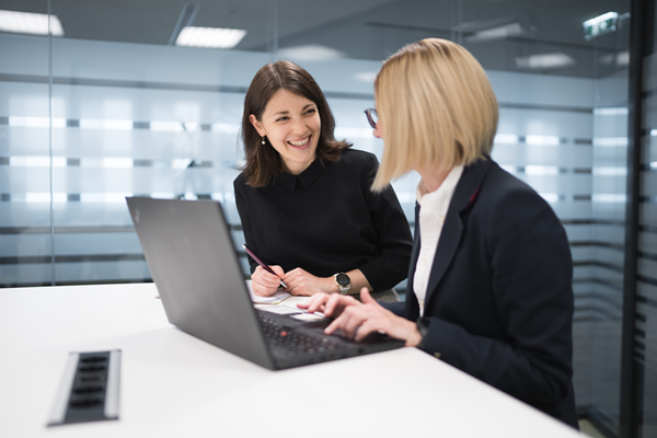 Zwei Mitarbeiterinnen sitzen mit einem Laptop im Besprechungsraum und lachen.