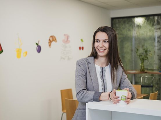 Жена в сив костюм държи чаша, стоейки в модерен офис с илюстрации на храни по стените и столове на заден план. Тази обстановка създава уютна и креативна атмосфера, която насърчава вдъхновението и взаимодействието.