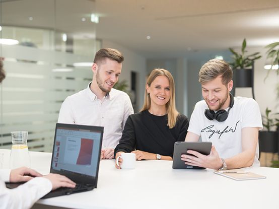 Четирима души в офис – трима стоят и се усмихват на таблет, а един работи на лаптоп. Светлата и модерна обстановка подчертава екипната работа и иновациите.
