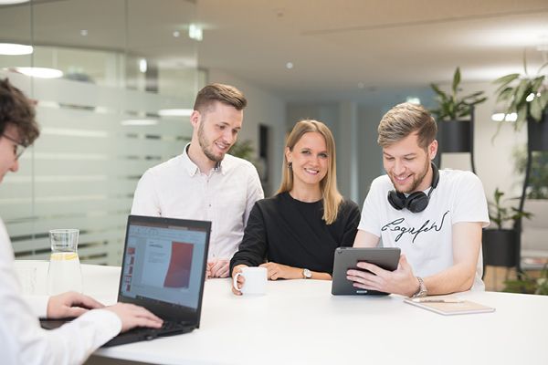 Четирима души в офис – трима стоят и се усмихват на таблет, а един работи на лаптоп. Светлата и модерна обстановка подчертава екипната работа и иновациите.