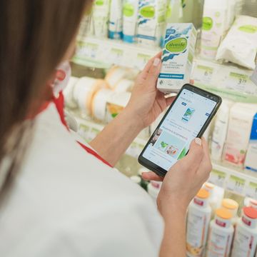 Човек в магазина сравнява козметичен продукт с информация на смартфона си. На заден план се виждат рафтове с разнообразни продукти, което подчертава богатия асортимент и възможността за информирано пазаруване.