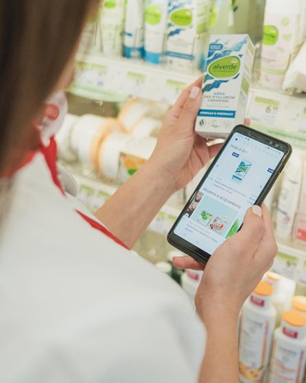 Човек в магазина сравнява козметичен продукт с информация на смартфона си. На заден план се виждат рафтове с разнообразни продукти, което подчертава богатия асортимент и възможността за информирано пазаруване.